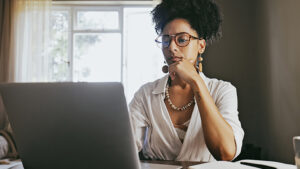 A woman in her 30s sits at a table in her home looking at her laptop as she reads about saving for retirement and how to start saving young.