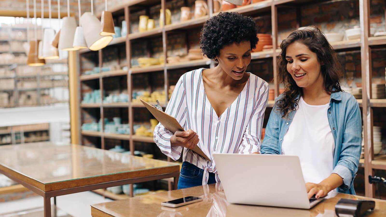 Two female colleagues standing at a table looking at a laptop.