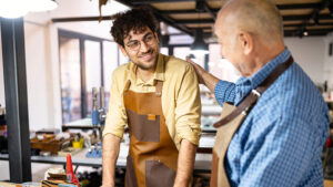 A father proudly looks at his son working alongside him in the workshop of their family business.