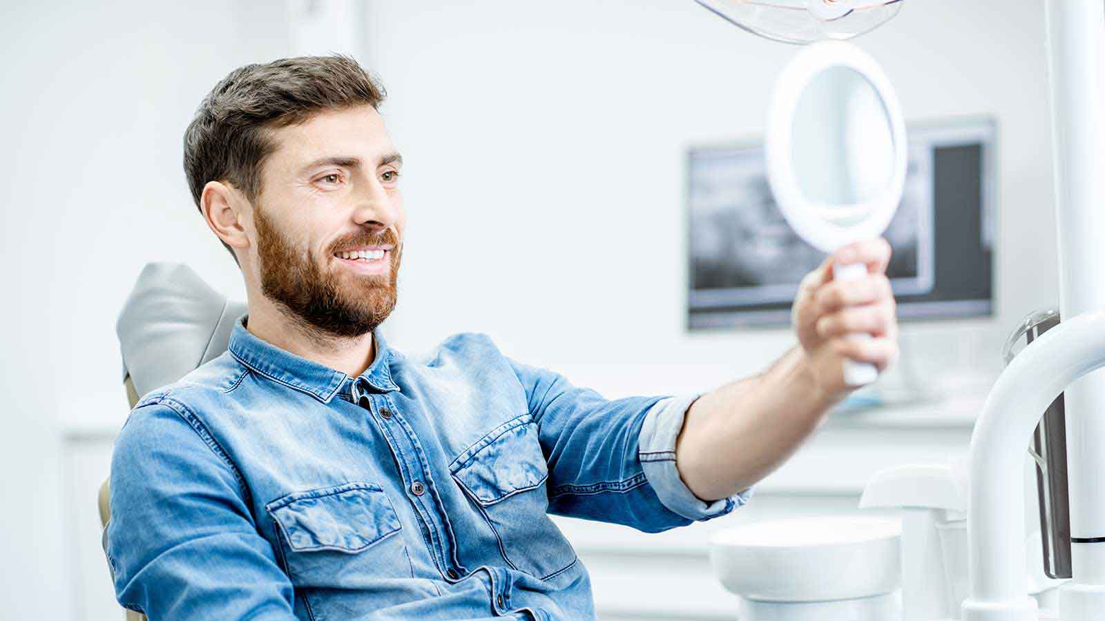 Man with beard smiling looking in mirror while sitting in dental chair.