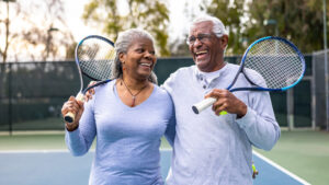 A husband and wife in their 60s walk off a paddleboard court after finishing a game with their arms around each other smiling.