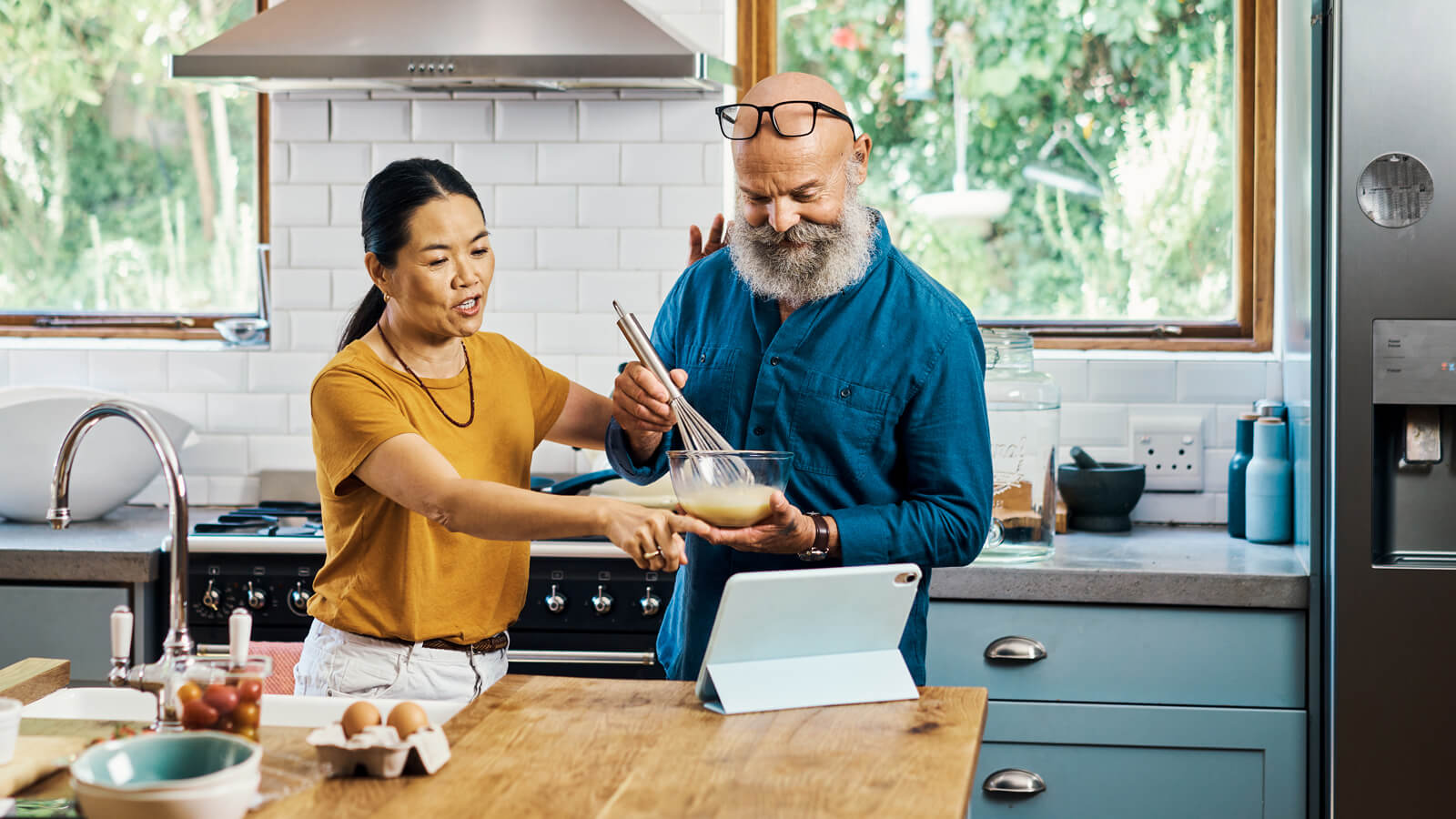 A husband and wife in their late 50s read a new recipe from their tablet on the kitchen counter while they cook together.