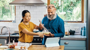 A husband and wife in their late 50s read a new recipe from their tablet on the kitchen counter while they cook together.