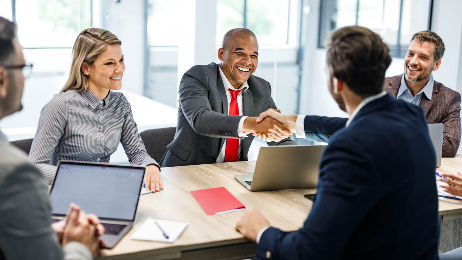 A group of four happy financial professionals sit around a conference table and shake hands with their new RIA representative after business disruption difficulties.