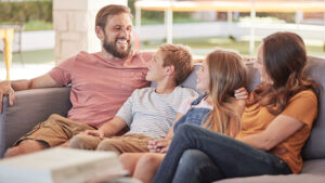 A dad and mom sit on either side of their two kids on an outdoor couch, the whole family smiling happily on a beautiful, sunny day.