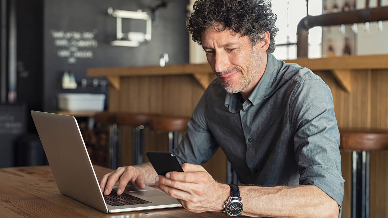 A man sits in a coffee shop using his laptop and cellphone to access personal information. 