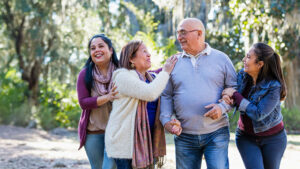 A family of four, two parents and their two adult daughters, are taking a walk on a sunny, fall day through a wooded park, laughing and having a fun afternoon together.