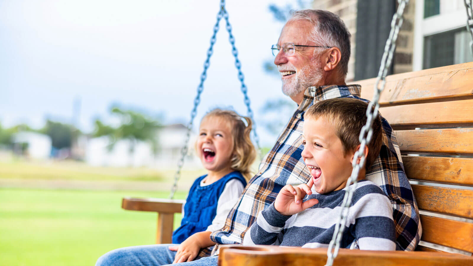 A retired grandfather sits on a front porch swing with his two young grandchildren on either side of him, and all three of them are smiling.