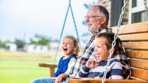 A retired grandfather sits on a front porch swing with his two young grandchildren on either side of him, and all three of them are smiling.