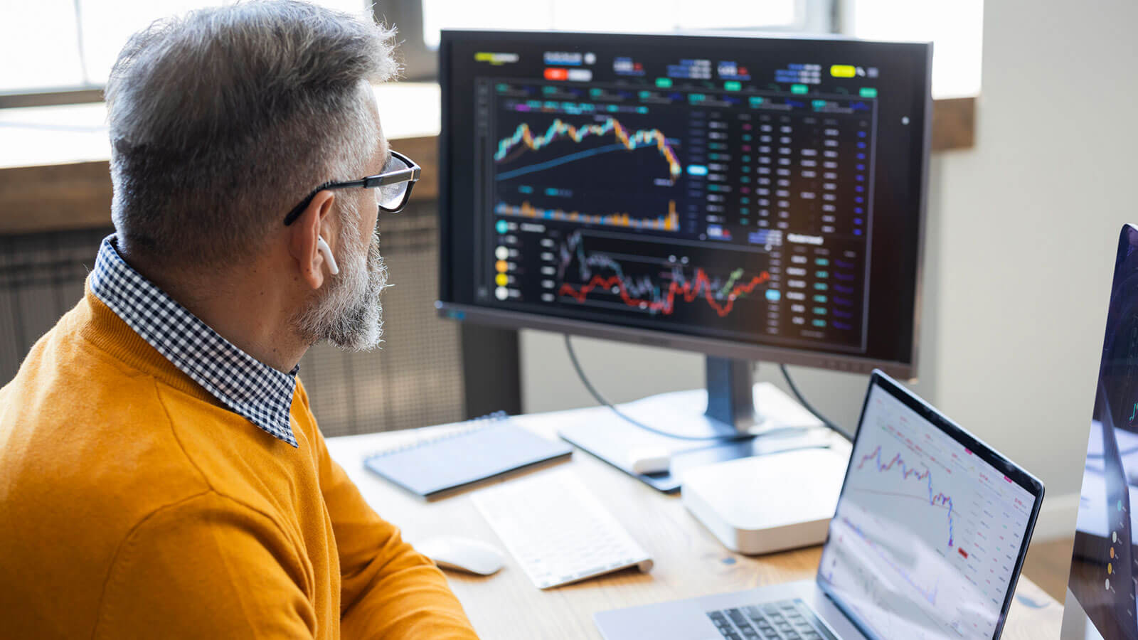 A man in his 50s wearing glasses sits at his desk reviewing stock market information on a large monitor screen and his laptop and thinks about strategies for surviving market turbulence.